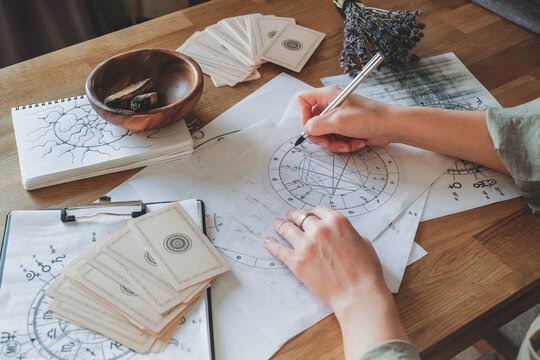Natal chart: an astrologer’s hands drawing a birth chart wheel on paper surrounded by notebooks and cards